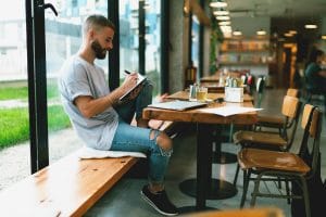 man writing in book in coffee shop with laptop on table