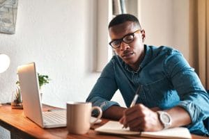 man taking notes with pen and paper while working on laptop